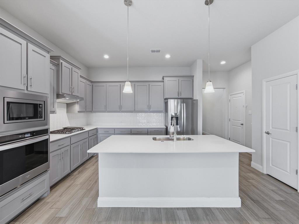 1121 Knightsbridge Road Argyle, TX 76226 - Photo 5 of 39 Kitchen with gray shaker-style cabinetry, stainless steel appliances, a center island featuring a sink, pendant lighting, and wood-finish flooring