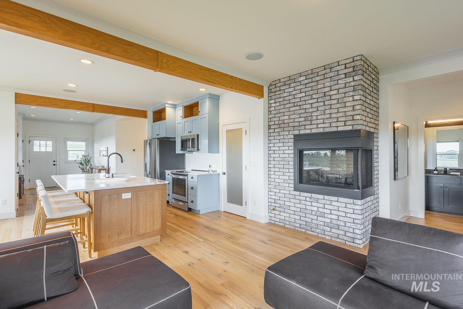 11171 Pump Road Nampa, ID 83686 - Photo 16 of 50 Open-concept interior featuring light wood-finish flooring, a two-sided brick fireplace with a black metal insert, and exposed wood ceiling beams