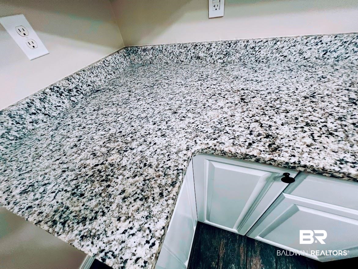 266 Ridgewood Drive Daphne, AL 36526 - Photo 9 of 34 Granite-look countertop with a built-in backsplash, complementing white cabinetry with dark hardware, and a durable wood-finish flooring