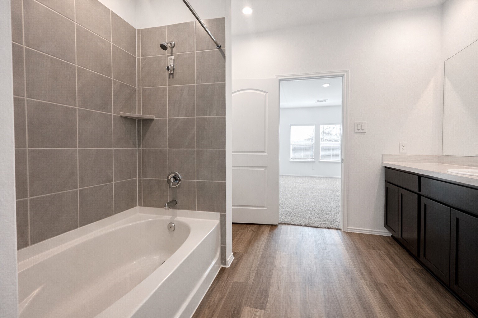1739 Royal Rim Drive Houston, TX 77014 - Photo 14 of 20 This bathroom features a modern design with a spacious bathtub surrounded by sleek gray tiles, a large mirror above a dark wood vanity, and wood-look flooring. It opens to a bright, carpeted bedroom.