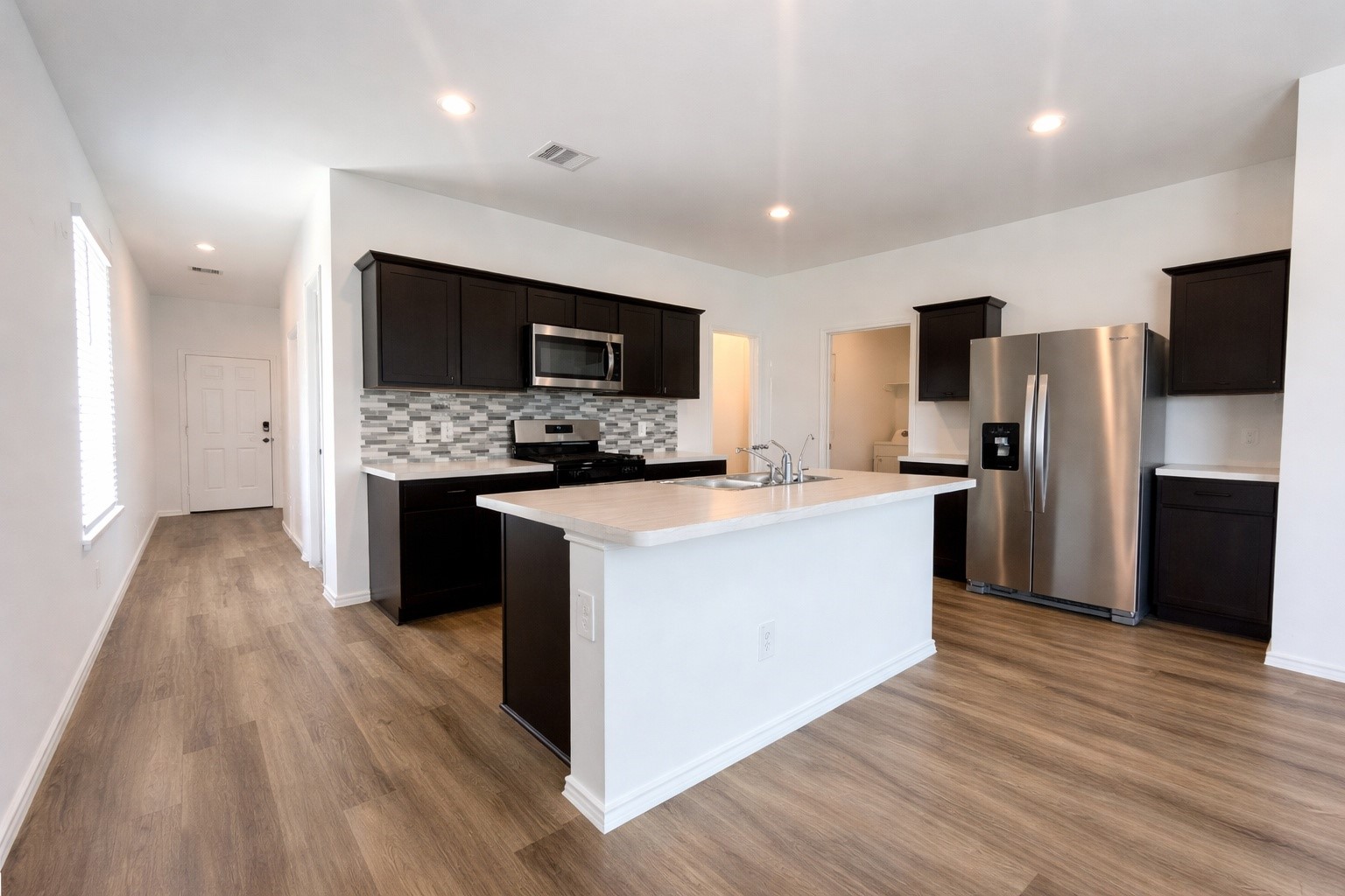 1739 Royal Rim Drive Houston, TX 77014 - Photo 4 of 20 A modern kitchen with sleek dark cabinetry, stainless steel appliances, and a stylish tile backsplash. The open layout includes a spacious island with a sink, set against a backdrop of light wood flooring and ample natural light.