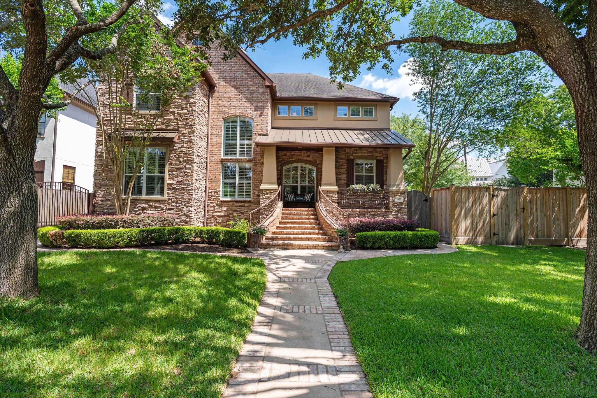 Welcome to 3822 Merrick St. A grand first impression awaits from the street, where a brick-bordered concrete walkway winds through a lush, manicured lawn framed by mature oak trees. The two-story façade combines rich brick and stacked stone with a standing seam metal roof over a welcoming front porch, arched entryway, and wrought iron stair railings. A cedar privacy fence with a gated entry runs along the side of the property.

