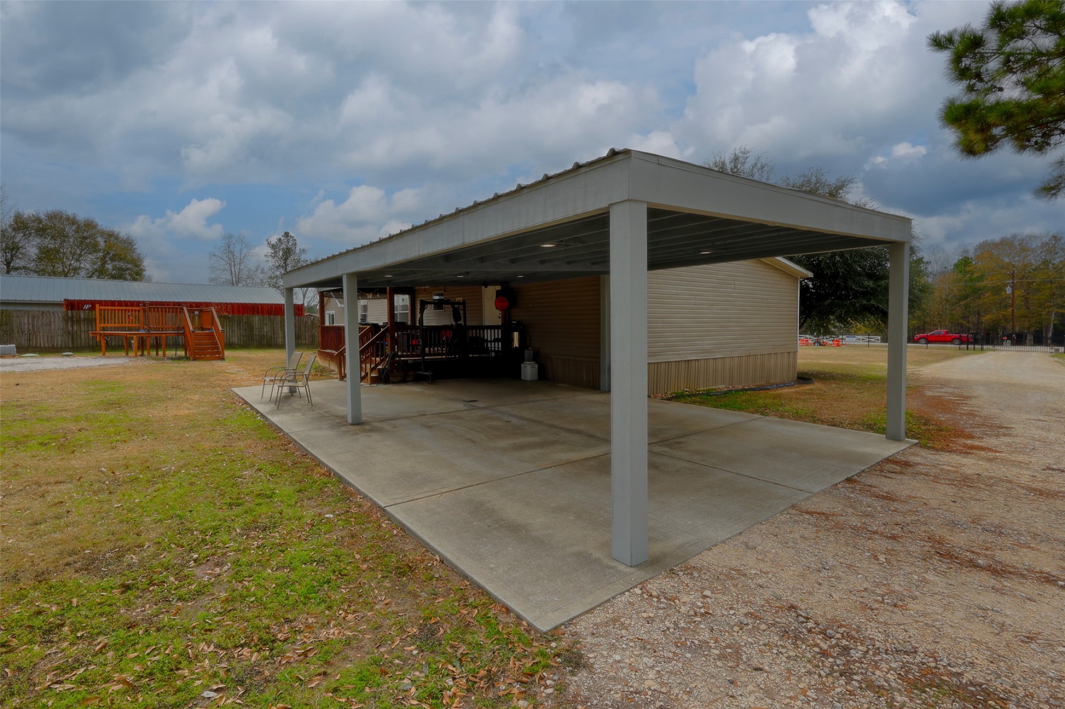 2373 Crockett Martin Road Conroe, TX 77306 - Photo 5 of 34 This photo features a covered outdoor area with a concrete floor, adjacent to a house. The space is suitable for parking or outdoor activities. In the background, there's a play structure and a fenced yard, ideal for family recreation. The surrounding area is spacious, with some trees and open sky visible.