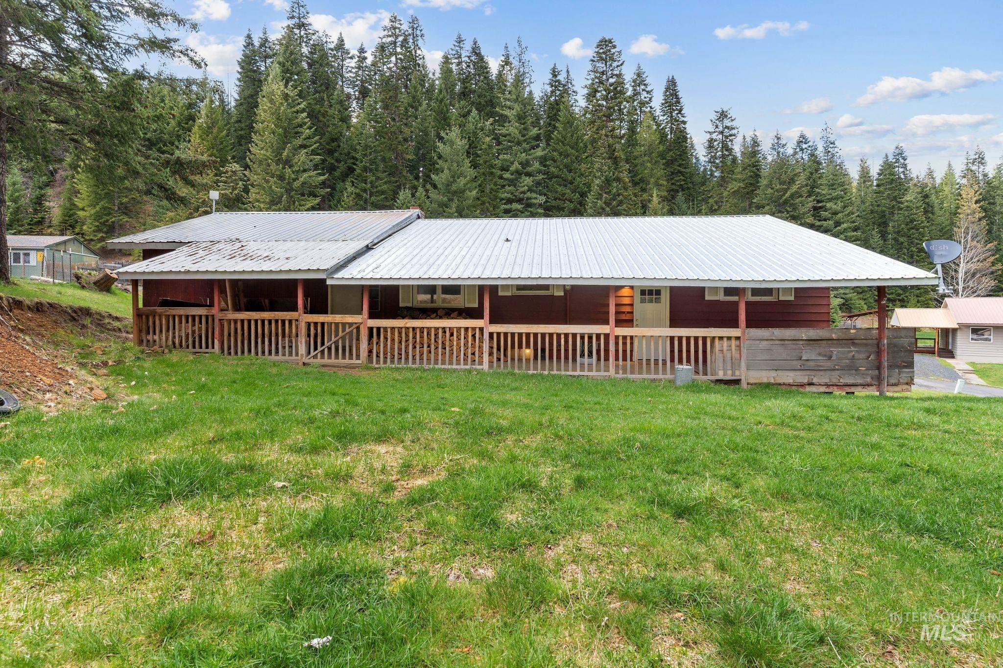 145 Timberline Drive Pierce, ID 83546 - Photo 36 of 39 Single-story red-sided dwelling featuring an extended covered front porch with a wood railing and posts, set against a backdrop of mature evergreen trees