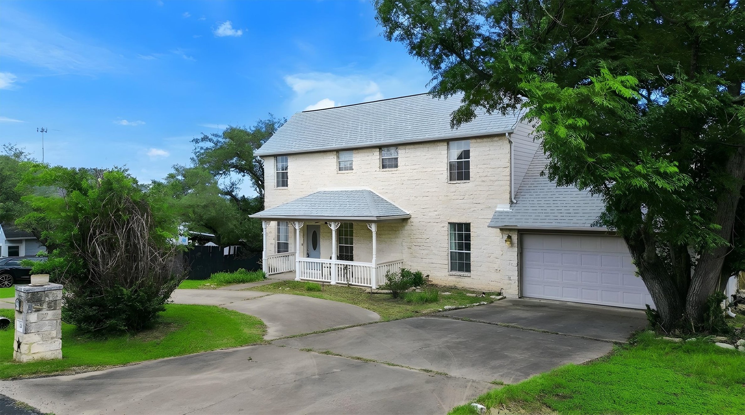 Two-story residence featuring light-colored exterior stonework, a covered front porch with white railings, a gray shingle roof, an attached garage, and mature landscaping