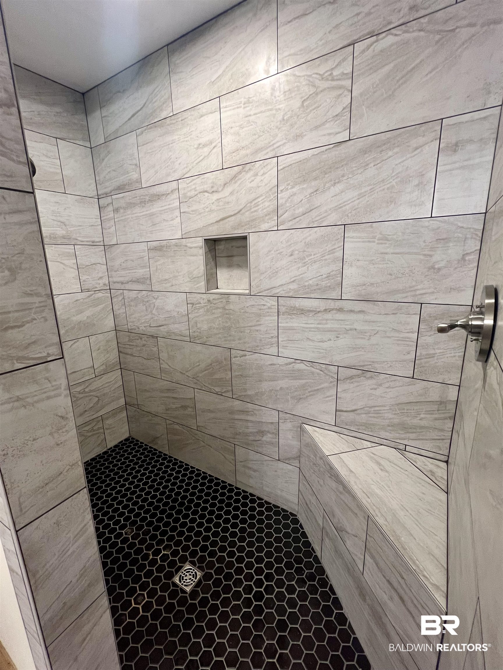29685 Gardner Road Elberta, AL 36530 - Photo 12 of 16 Walk-in shower featuring light-toned rectangular wall tiles, a built-in shower niche, a brushed metal shower fixture, and dark hexagonal floor tiles with a central drain