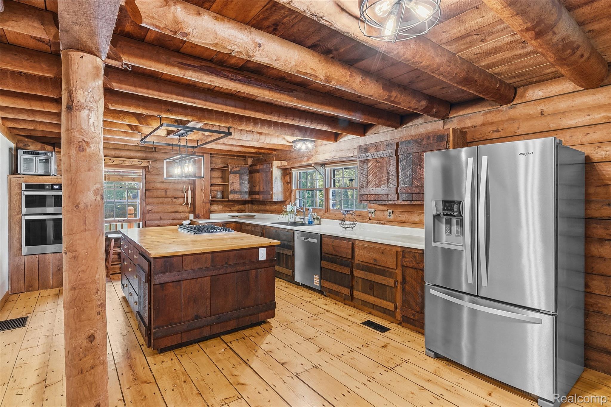 3656 Stanley Road Columbiaville, MI 48421 - Photo 12 of 65 Kitchen featuring wood-finish flooring, exposed wood beam ceiling, wood plank walls, a central island with wood countertop and cooktop, and stainless steel appliances including a refrigerator, dishwasher, and built-in double oven