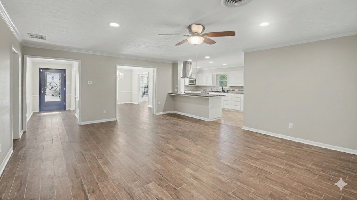 1431 Walnut Lane Houston, TX 77339 - Photo 31 of 46 This photo showcases a spacious open-concept living area with wood-look flooring, neutral walls, and recessed lighting. It features a modern kitchen with a central island and stainless steel appliances, visible through a wide entryway. A ceiling fan adds comfort, and a doorway leads to a well-lit entryway with a decorative glass door.