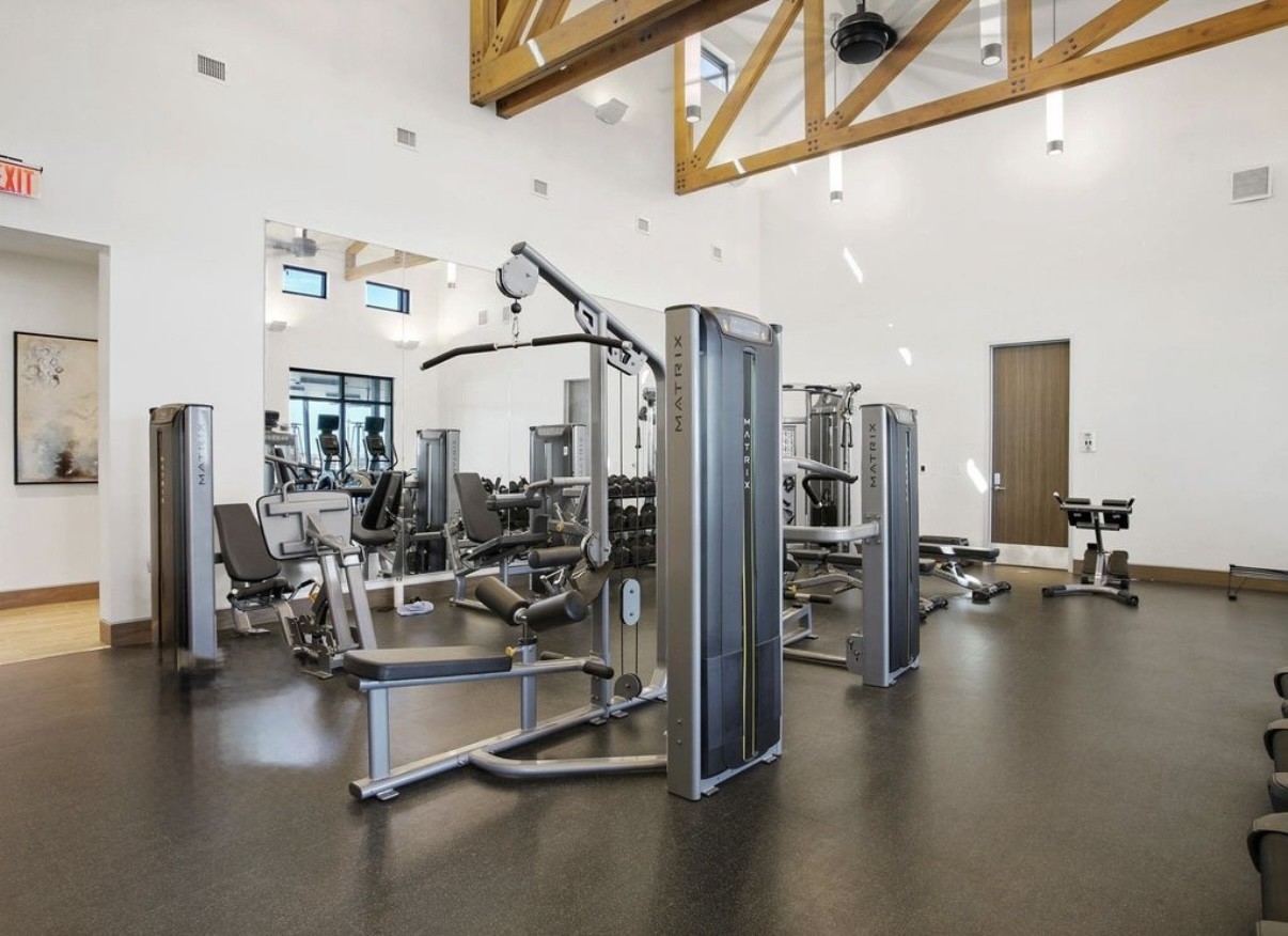 3006 Floating Barque Drive Santa Fe, TX 77510 - Photo 37 of 37 This spacious home gym features modern exercise equipment, high ceilings with exposed wooden beams, large mirrors, and ample natural light, creating a bright and inviting workout environment.