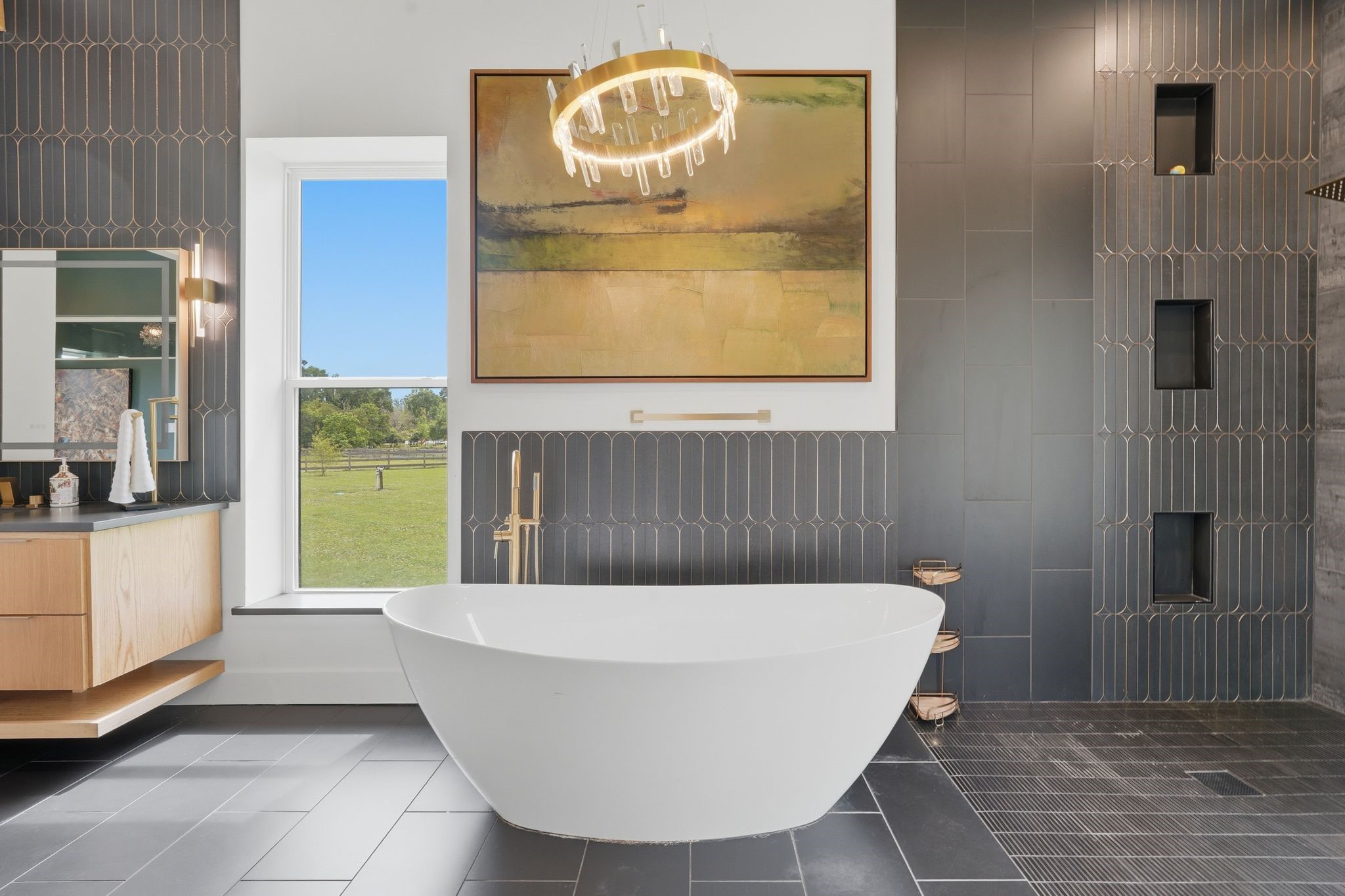 2743 Bobville Road Montgomery, TX 77316 - Photo 31 of 50 This modern bathroom features a sleek freestanding tub, elegant dark tilework, and a large window offering scenic views. A contemporary chandelier and wall art add a touch of luxury, while ample storage and stylish fixtures complete the space.