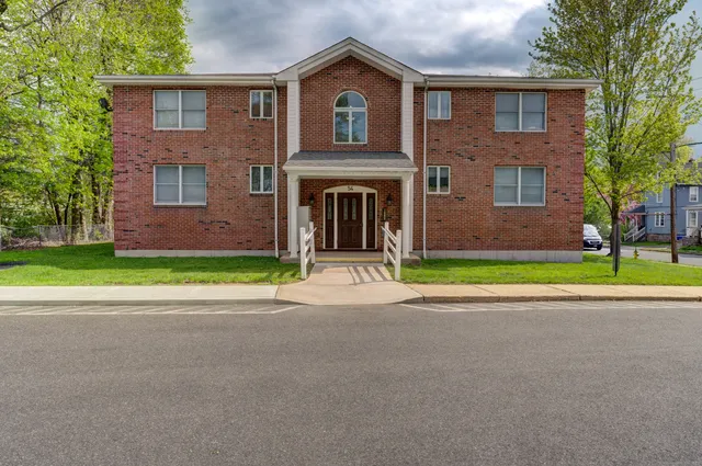 a view of a brick house with a yard plants and large tree