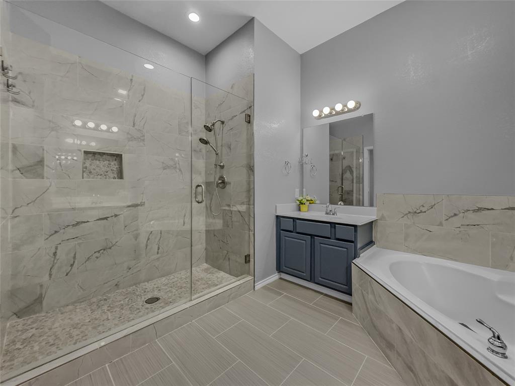 3008 Devlin Ridge Drive Plano, TX 75025 - Photo 11 of 24 Spacious bathroom featuring a glass-enclosed shower with tile surround and integrated niche, a separate soaking tub with tile surround, a single vanity with a dark wood cabinet and light countertop, a wall-mounted mirror, and neutral-toned tile flooring