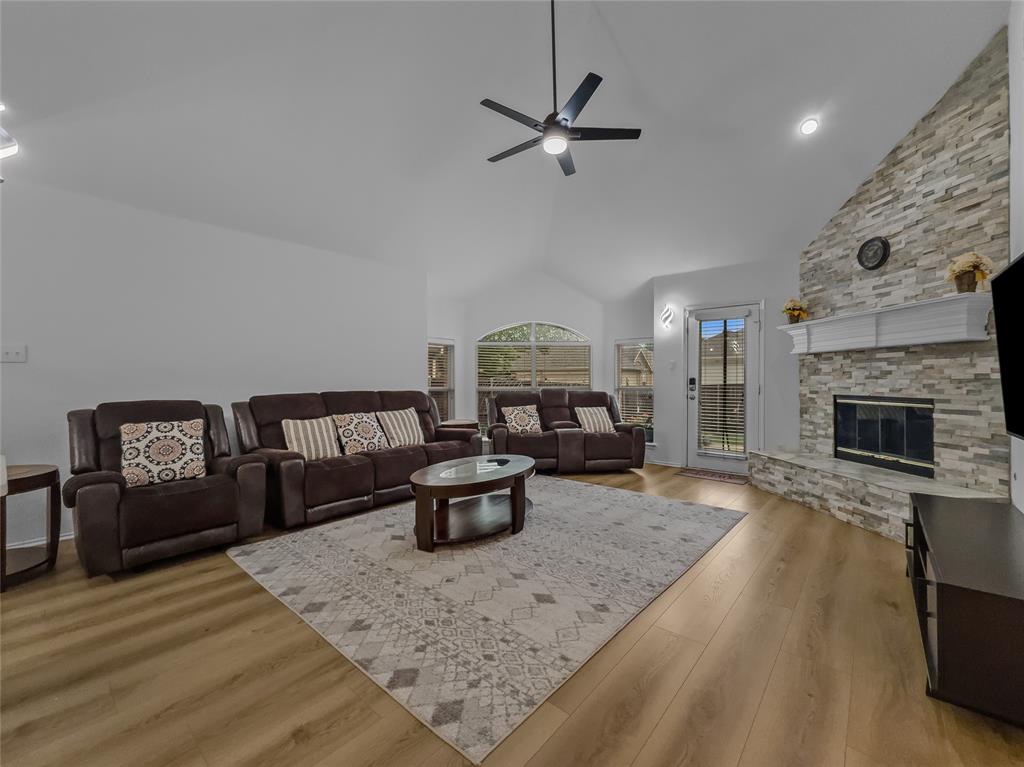 3008 Devlin Ridge Drive Plano, TX 75025 - Photo 9 of 24 Spacious living area featuring a vaulted ceiling with a ceiling fan, a stone-faced fireplace with a white mantel, and wood-finish flooring