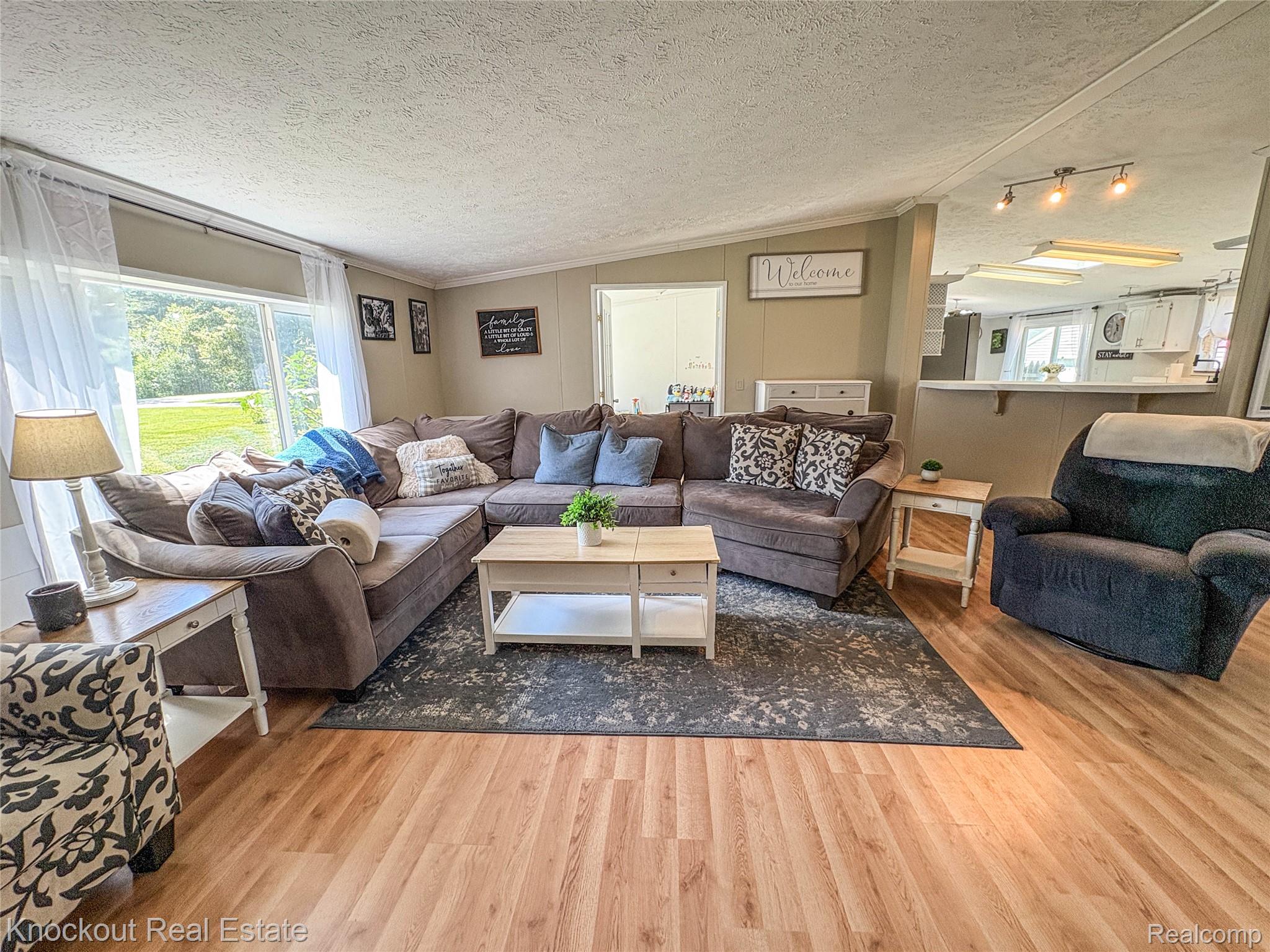 7760 Bell Road Birch Run, MI 48415 - Photo 6 of 68 Spacious living area featuring wood-finish flooring, a large picture window, track lighting, and a partial wall with a breakfast bar opening to the kitchen
