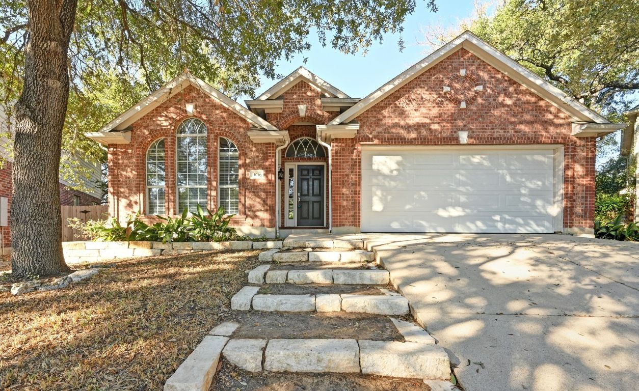 Welcome Home! Charming red brick façade accented by arched windows and a classic black front door create timeless curb appeal. Mature trees provide shade and a welcoming presence along the stone walkway.