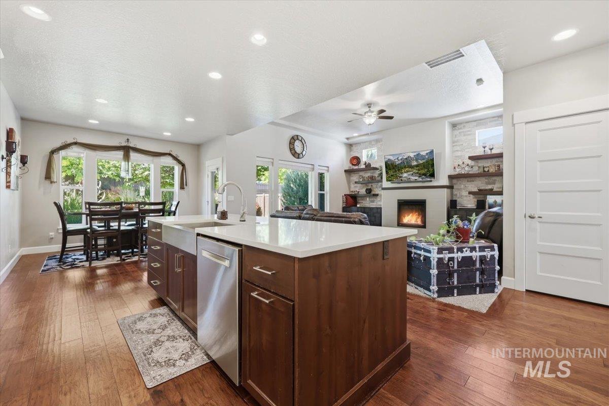 1303 West Brando Street Meridian, ID 83646 - Photo 14 of 49 Open-concept living space featuring wood-finish flooring, a kitchen island with a farmhouse sink and stainless steel dishwasher, and a living area with a built-in fireplace and stacked stone surround