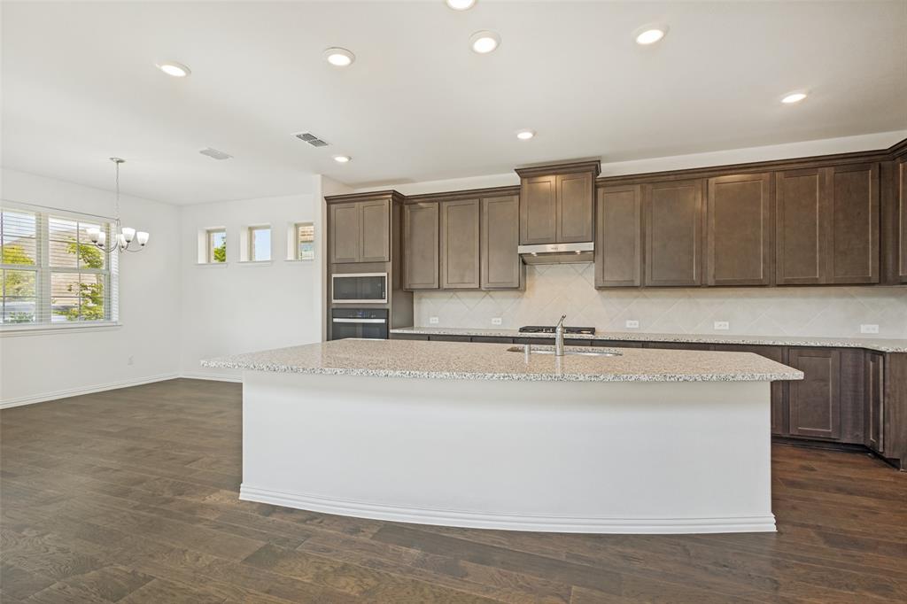 1448 Sun Gdn Way Justin, TX 76247 - Photo 5 of 15 Kitchen featuring a large island with a light-toned countertop, dark wood-finish cabinetry, built-in double ovens, and a tile backsplash