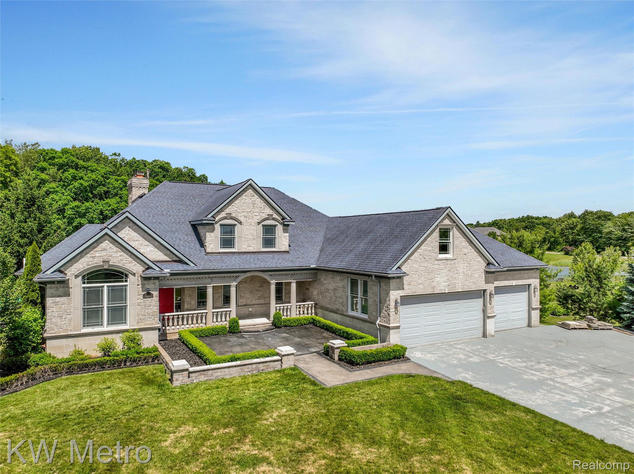 65884 Windrose Court Ray, MI 48096 - Photo 3 of 82 Stone facade residence featuring a covered front porch with column supports, a prominent red entry door, and a multi-car garage