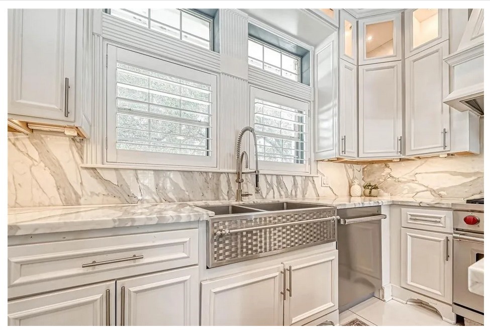 263 Wood Loop Street Houston, TX 77015 - Photo 19 of 45 This kitchen features elegant white cabinetry, a marble backsplash, and countertops. It includes a deep farmhouse sink, modern fixtures, and ample natural light from large windows with shutters, creating a bright and sophisticated space.