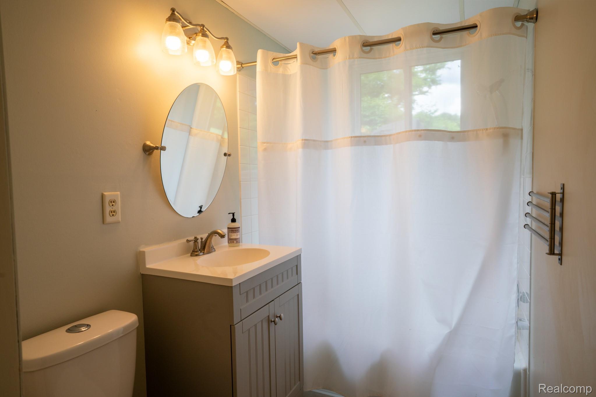 7333 Toma Road Dexter, MI 48130 - Photo 36 of 45 Bathroom featuring a single vanity with a gray cabinet and an integrated sink, an oval mirror, a three-light fixture, and a window with a shower curtain