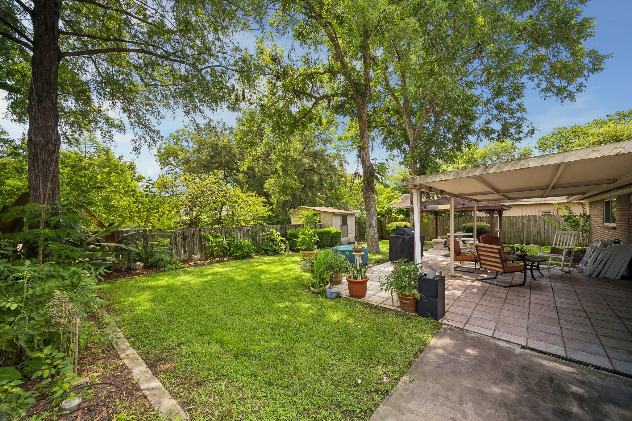 413 Mebane Lane Rosenberg, TX 77471 - Photo 29 of 30 This backyard features a lush green lawn surrounded by mature trees and a wooden fence for privacy. There's a spacious tiled patio with comfortable seating and a covered area, perfect for outdoor entertaining. There is additional yard space on the other side of the fence.