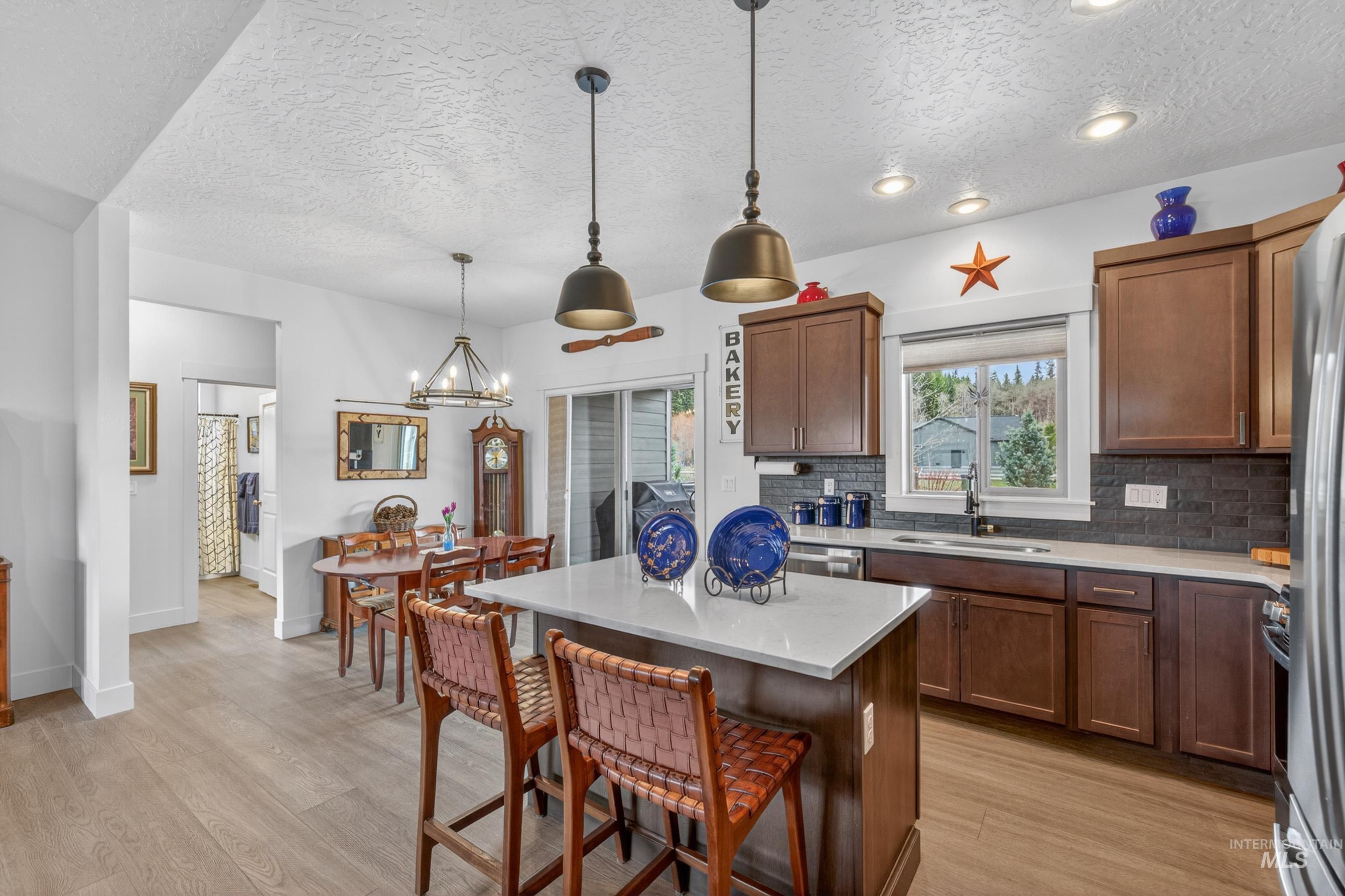 1126 Mo's Way McCall, ID 83638 - Photo 11 of 50 Open-concept kitchen featuring an island with a light-toned countertop and pendant lighting, wood-finish flooring, recessed lighting, brown cabinetry, and a dark tile backsplash