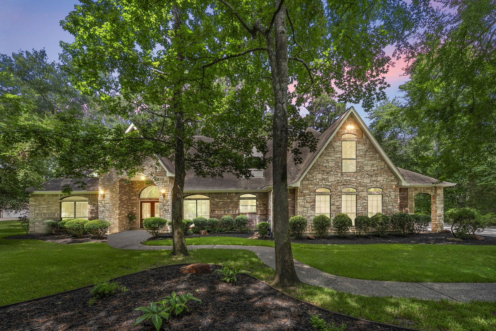 24311 Standing Oak Drive Spring, TX 77389 - Photo 2 of 42 Arrive home through a canopy of mature trees to a stately stone and brick façade that feels tucked away from the world. Soft exterior lighting and a winding walk create a welcoming approach for evenings with friends or quiet nights in.