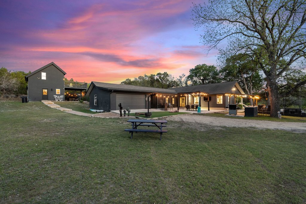 2800 River Road Wimberley, TX 78676 - Photo 5 of 38 Sprawling property featuring a multi-structure layout with dark-toned exteriors, a covered patio with string lighting, a detached garage, and a lush, expansive lawn