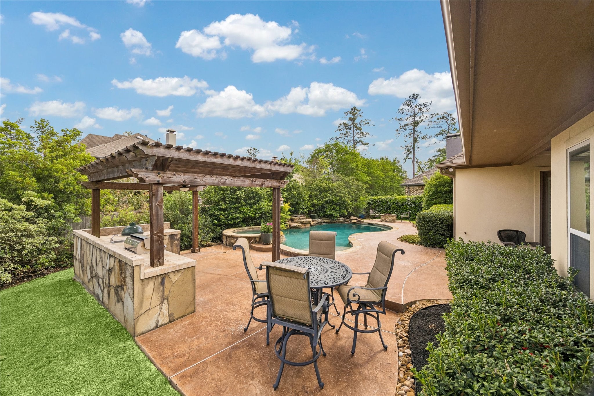 3 Tiburon Court Spring, TX 77389 - Photo 29 of 37 This backyard features a beautifully landscaped area with a swimming pool, a stone patio with outdoor dining set, and a wooden pergola housing a built-in grill, surrounded by lush greenery for privacy.