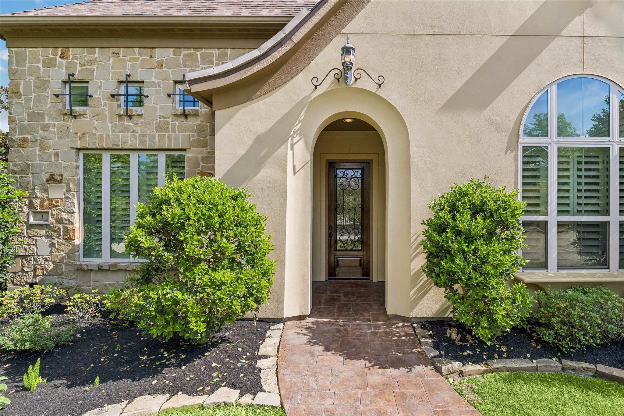 3 Tiburon Court Spring, TX 77389 - Photo 3 of 37 This photo showcases a charming home entrance with a stone and stucco façade, arched doorway, and decorative iron door. The landscaping features manicured bushes and a stone-bordered walkway, enhancing the home's curb appeal.