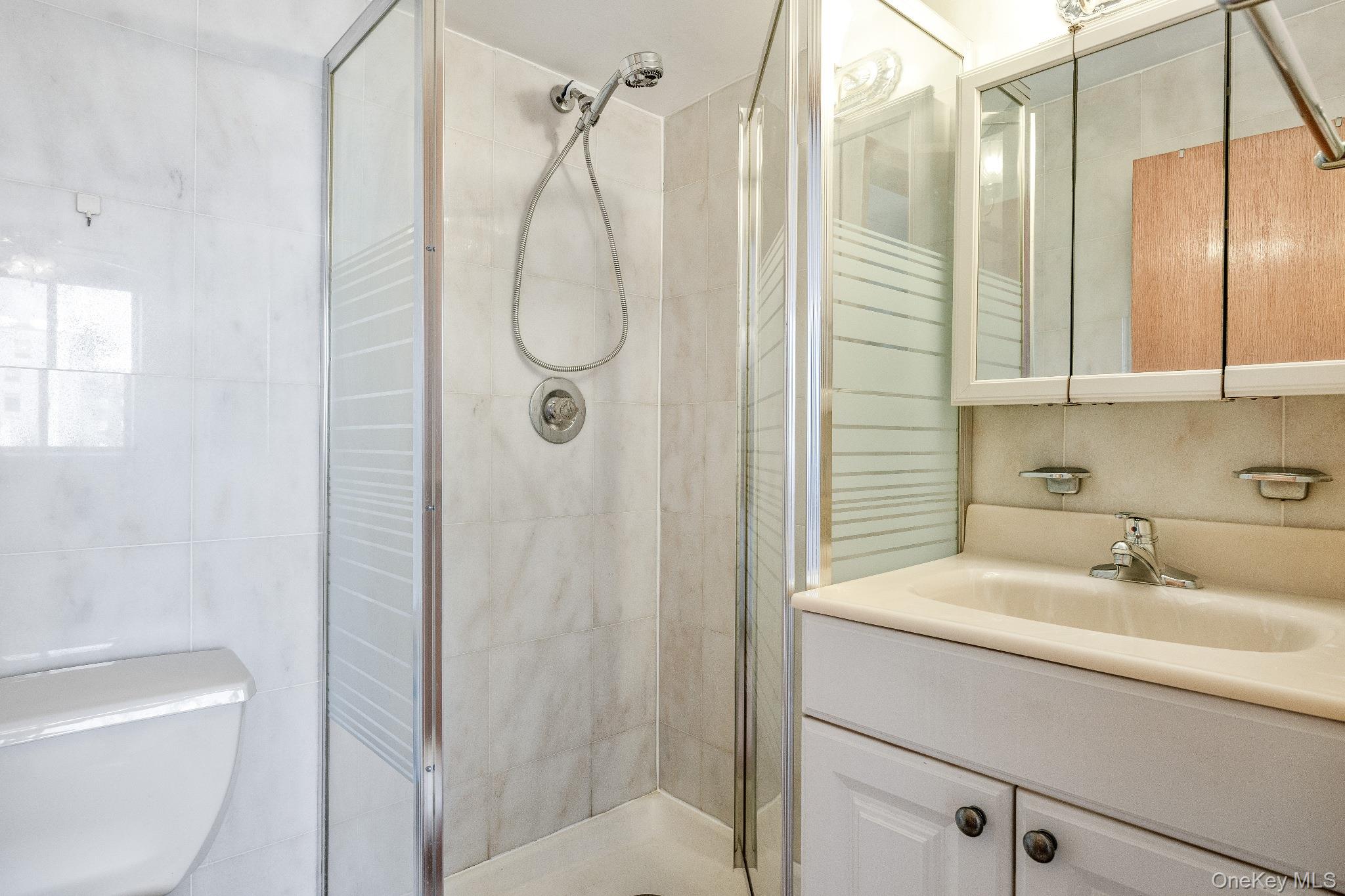 133-36 41st Road, Unit 8F Queens, NY 11355 - Photo 13 of 18 Second Bathroom / Shower – This bathroom features tiled walls, a glass-enclosed stand-up shower, and a vanity with integrated sink and mirrored medicine cabinet for convenient storage. The efficient layout provides a practical and functional space for daily use.