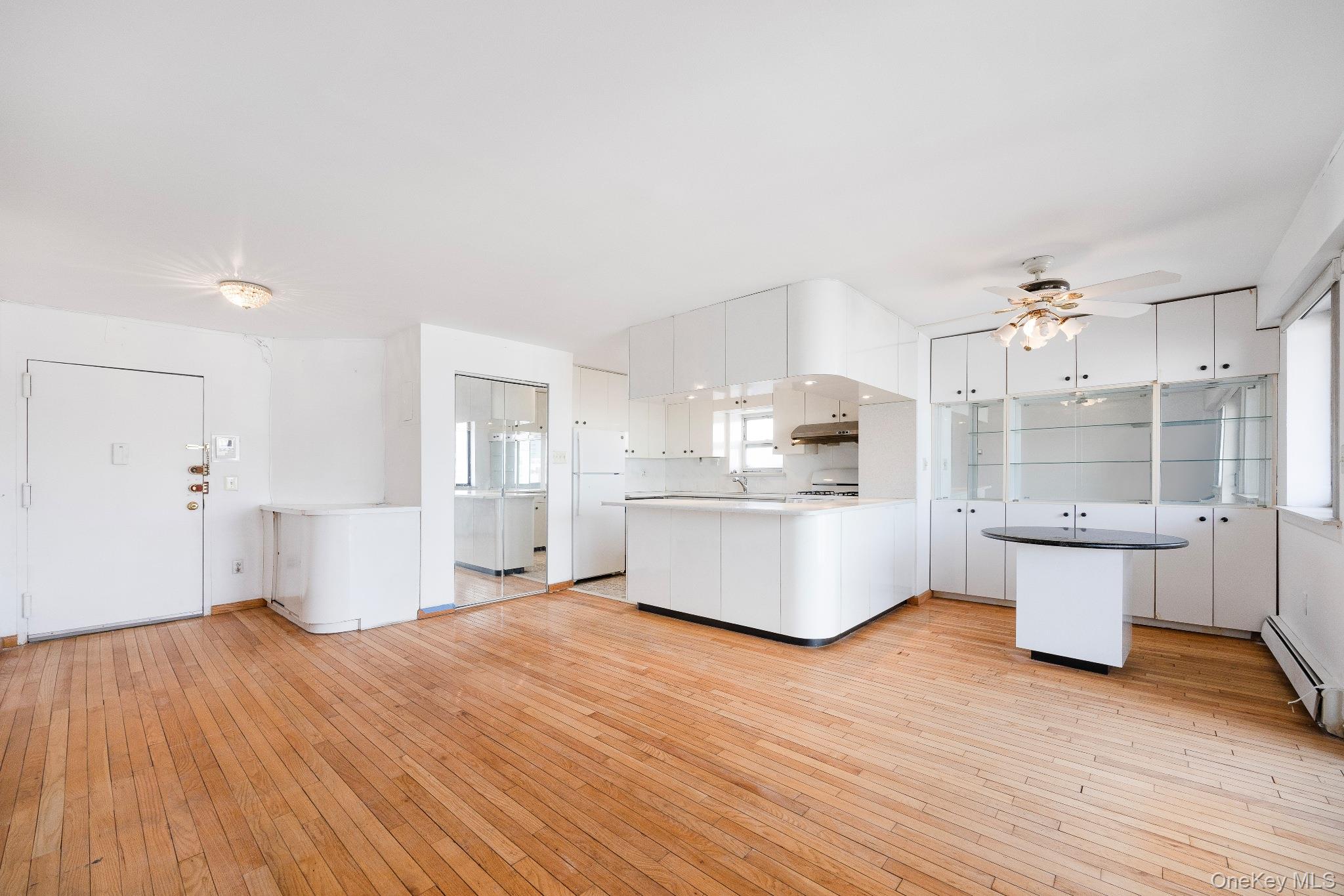 133-36 41st Road, Unit 8F Queens, NY 11355 - Photo 4 of 18 Living Room / Dining Area / Kitchen – This bright open-concept living and dining space features warm hardwood flooring and oversized windows that fill the home with natural light. The adjoining kitchen offers ample cabinetry, a convenient breakfast bar with seating, and a seamless layout ideal for everyday living and entertaining.