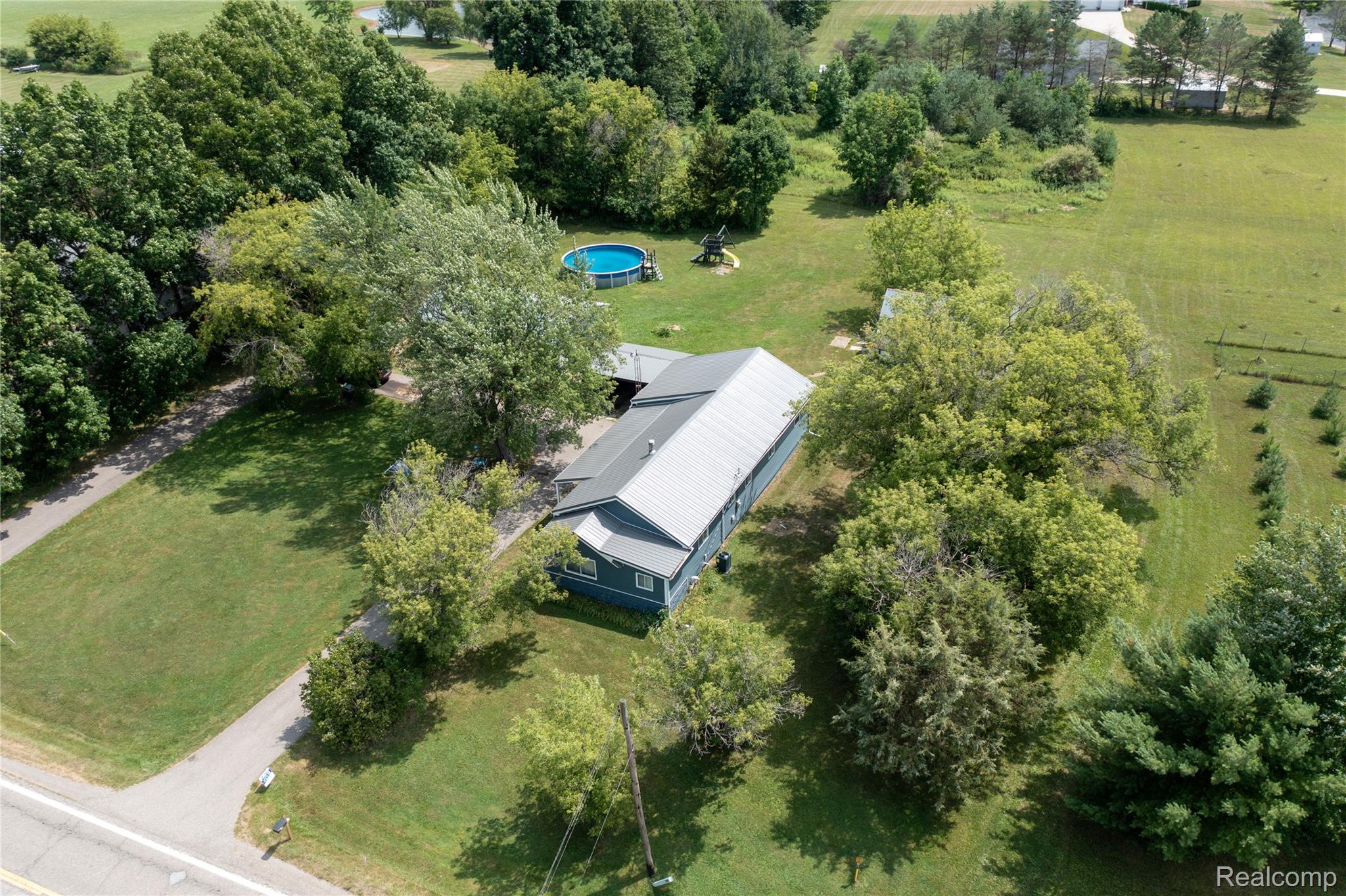 5761 Genesee Road Lapeer, MI 48446 - Photo 36 of 52 Aerial property view featuring a blue-sided residence with a gray metal roof, a paved driveway, mature trees, an above-ground pool, and expansive green spaces