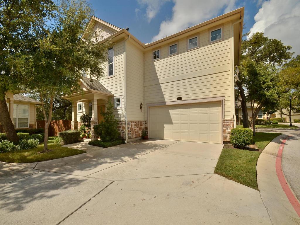 A cream-colored residence offers a welcoming front porch and an attached garage, nestled among mature trees and manicured shrubs.