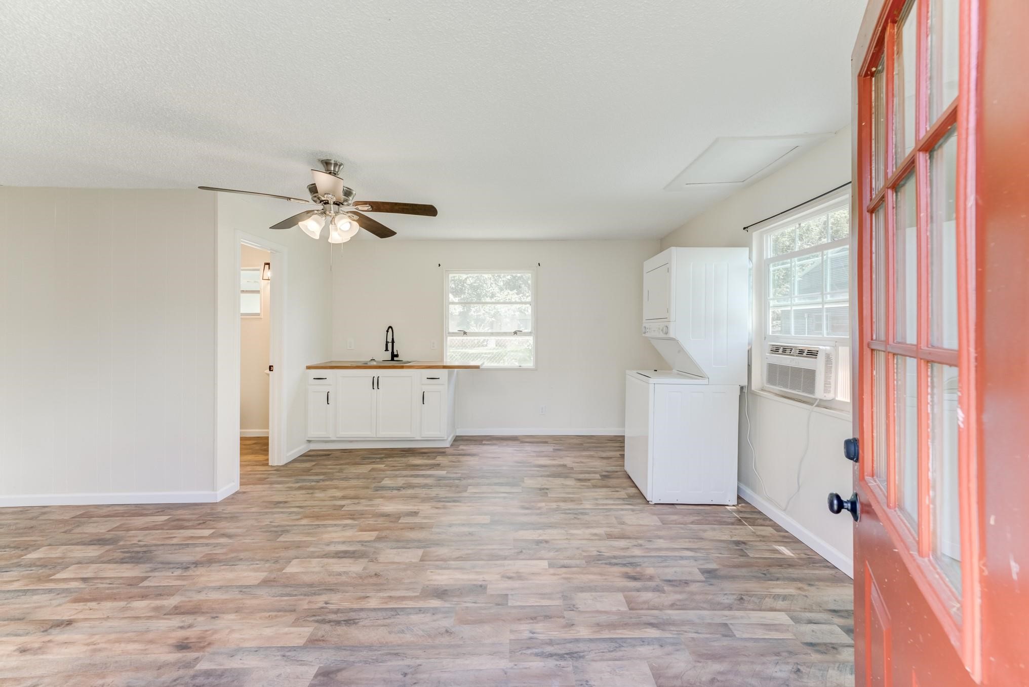 305 Yaupon Street Sweeny, TX 77480 - Photo 10 of 22 Bright and airy room with a ceiling fan, wood-look flooring, and a red door entrance. Features a small kitchenette with white cabinets, a black faucet, and a stackable washer and dryer. Large windows provide ample natural light.