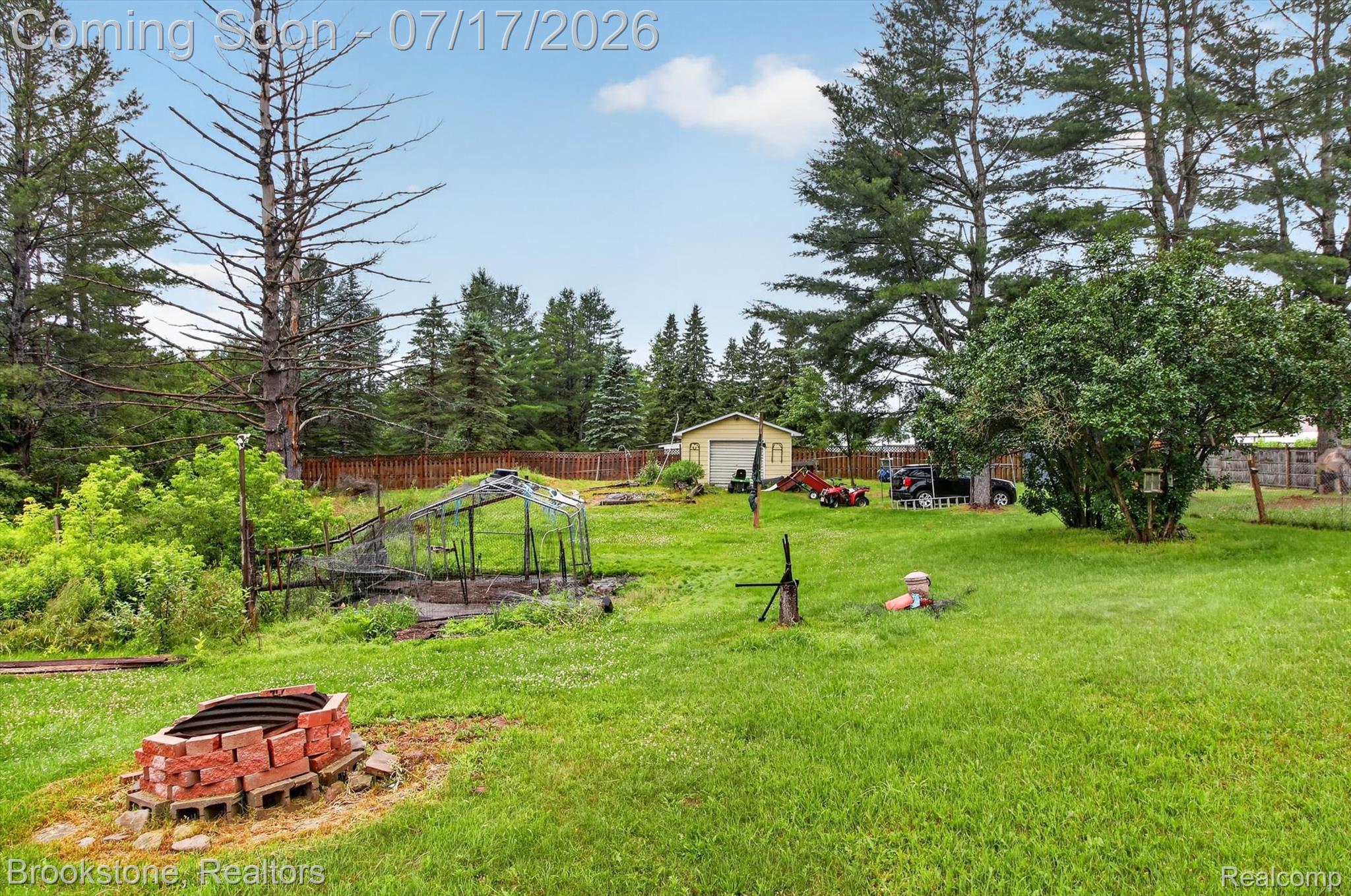 22709 Pleasant Valley Road Hillman, MI 49746 - Photo 31 of 42 Expansive grassy yard featuring a brick fire pit, a greenhouse structure, mature trees, and a detached storage shed with a roll-up door