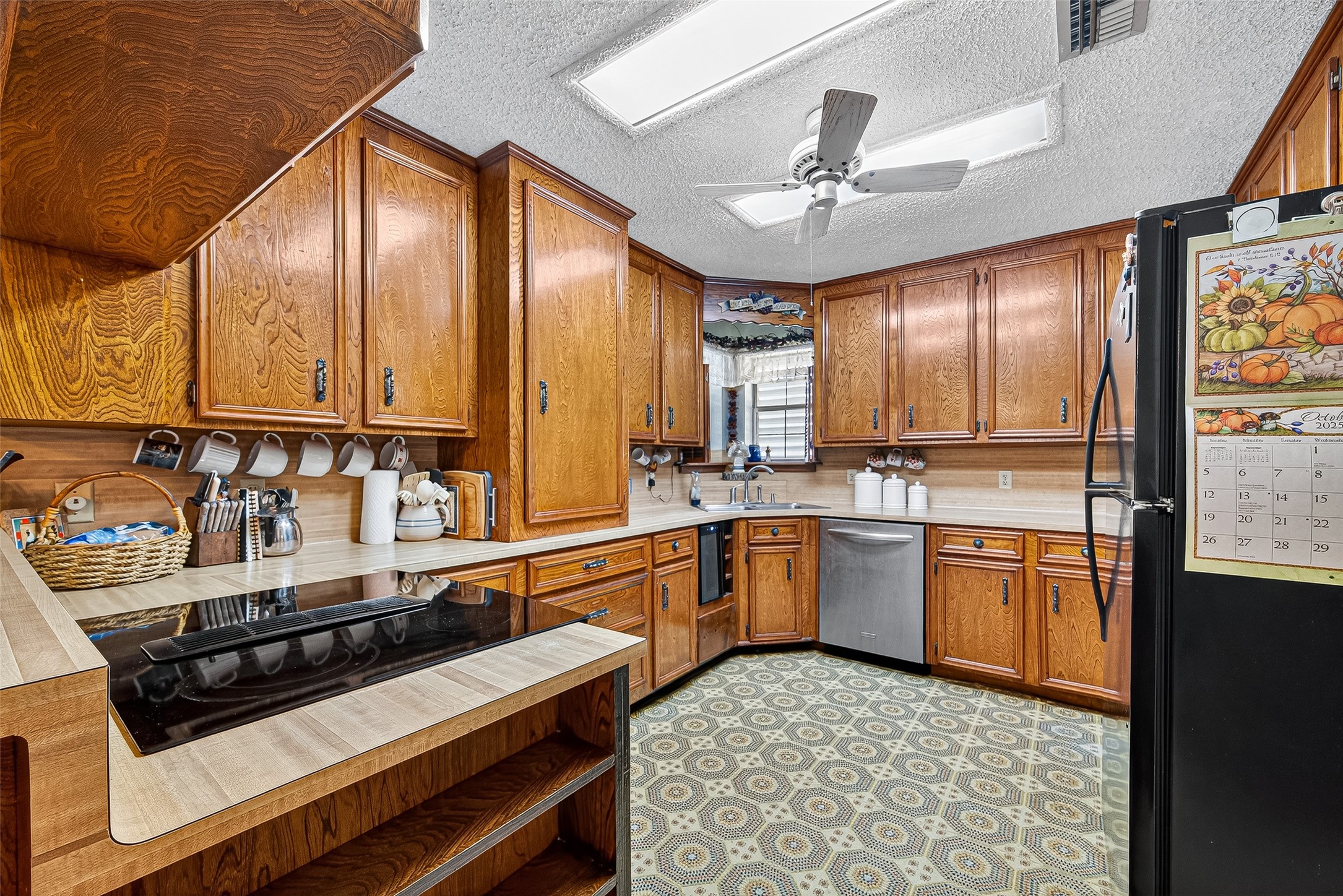 12021 7th 1/2 Street Santa Fe, TX 77510 - Photo 24 of 49 This kitchen features classic wooden cabinetry with ample storage, a modern cooktop, and a stainless steel dishwasher. It has a cozy atmosphere with patterned flooring, ceiling fan, and natural light from a window above the sink.
