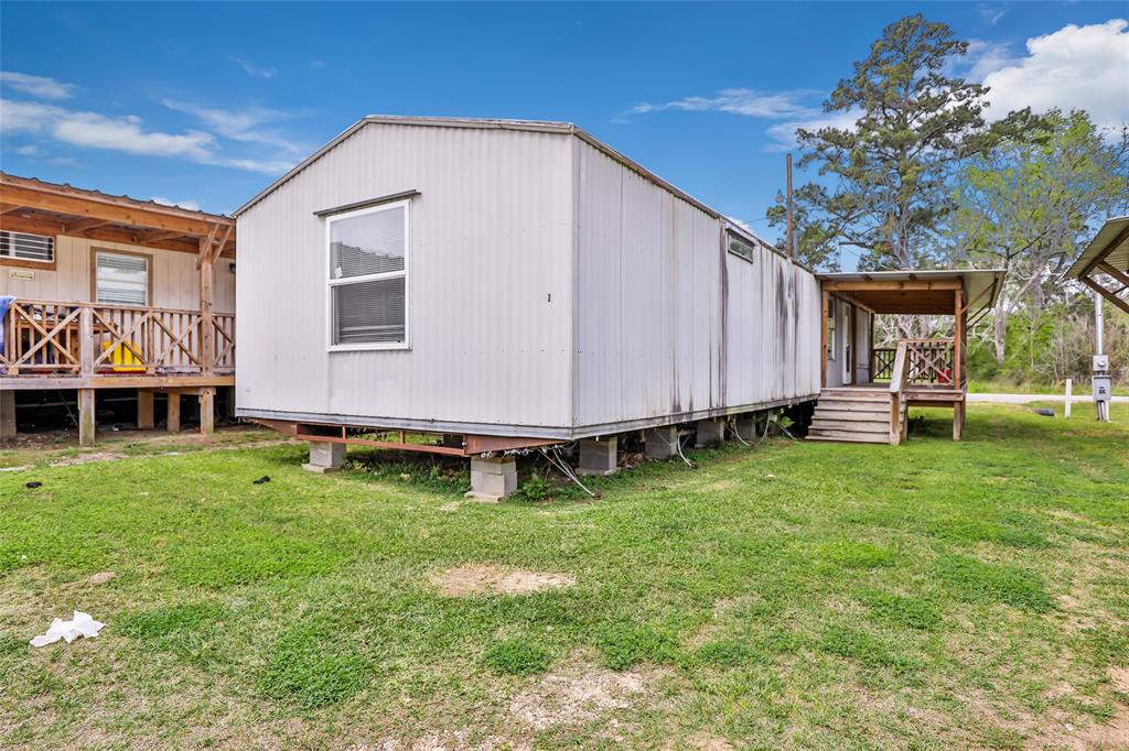 10821 Highway 150, Unit 18 Shepherd, TX 77371 - Photo 1 of 9 This photo shows a mobile home on a grassy lot, elevated on concrete blocks. It features a covered porch with wooden steps. The surrounding area is open with a few trees and adjacent structures.