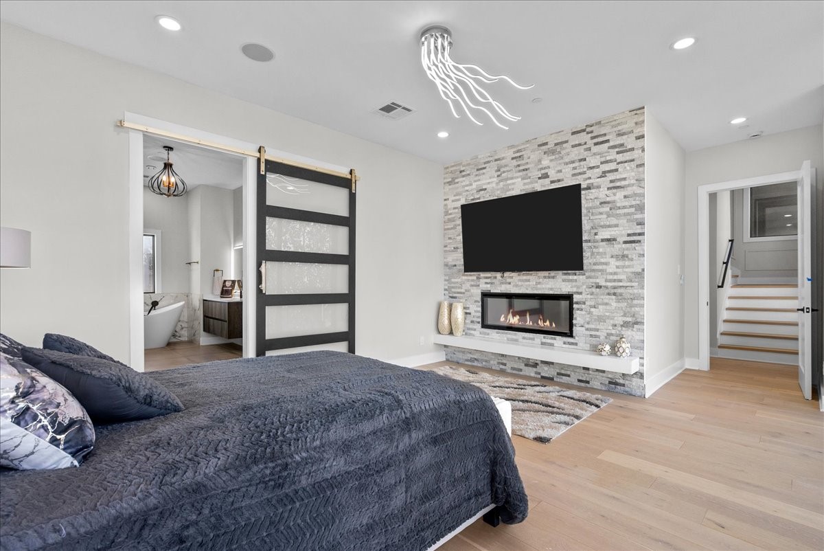 516 Reinerman Street Houston, TX 77007 - Photo 26 of 42 This modern bedroom features a cozy fireplace with a stone accent wall, a mounted TV, and sleek wood flooring. A sliding barn door leads to a luxurious bathroom with a soaking tub. Recessed lighting and a contemporary ceiling fixture add elegance.