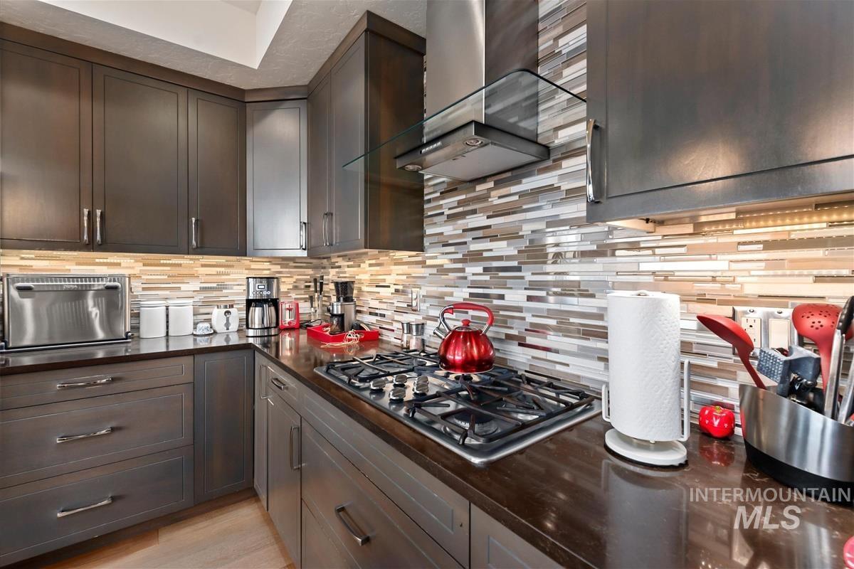 24398 Tombstone Ridge Court Middleton, ID 83644 - Photo 7 of 48 Dark wood-finish cabinetry with silver hardware, multi-tone tile backsplash, built-in gas range, stainless steel range hood with glass canopy, and dark countertops