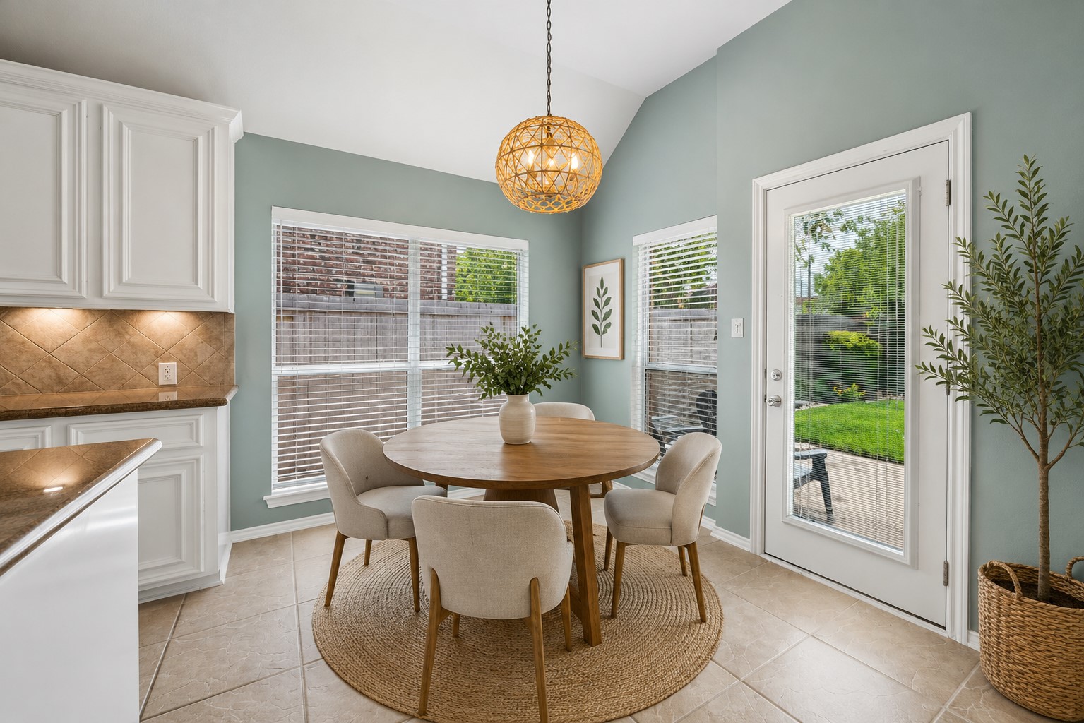 3118 Valley Court Manvel, TX 77578 - Photo 8 of 38 This inviting virtual staged dining area features a round wooden table with plush chairs, set on a cozy rug. It has a modern light fixture, ample natural light from large windows, and a door leading to a lush backyard. The soft blue walls complement the white cabinetry and tiled floor, creating a warm atmosphere.