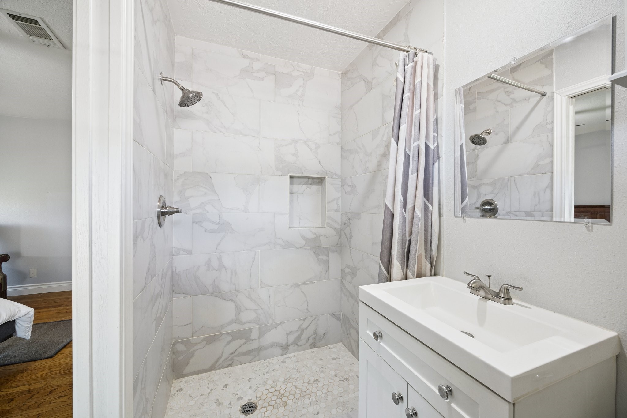 8911 Blankenship Drive Houston, TX 77080 - Photo 13 of 21 This bathroom features a sleek, modern design with elegant marble-patterned tiles in the shower, a convenient built-in niche, and a compact white vanity with a mirror above.