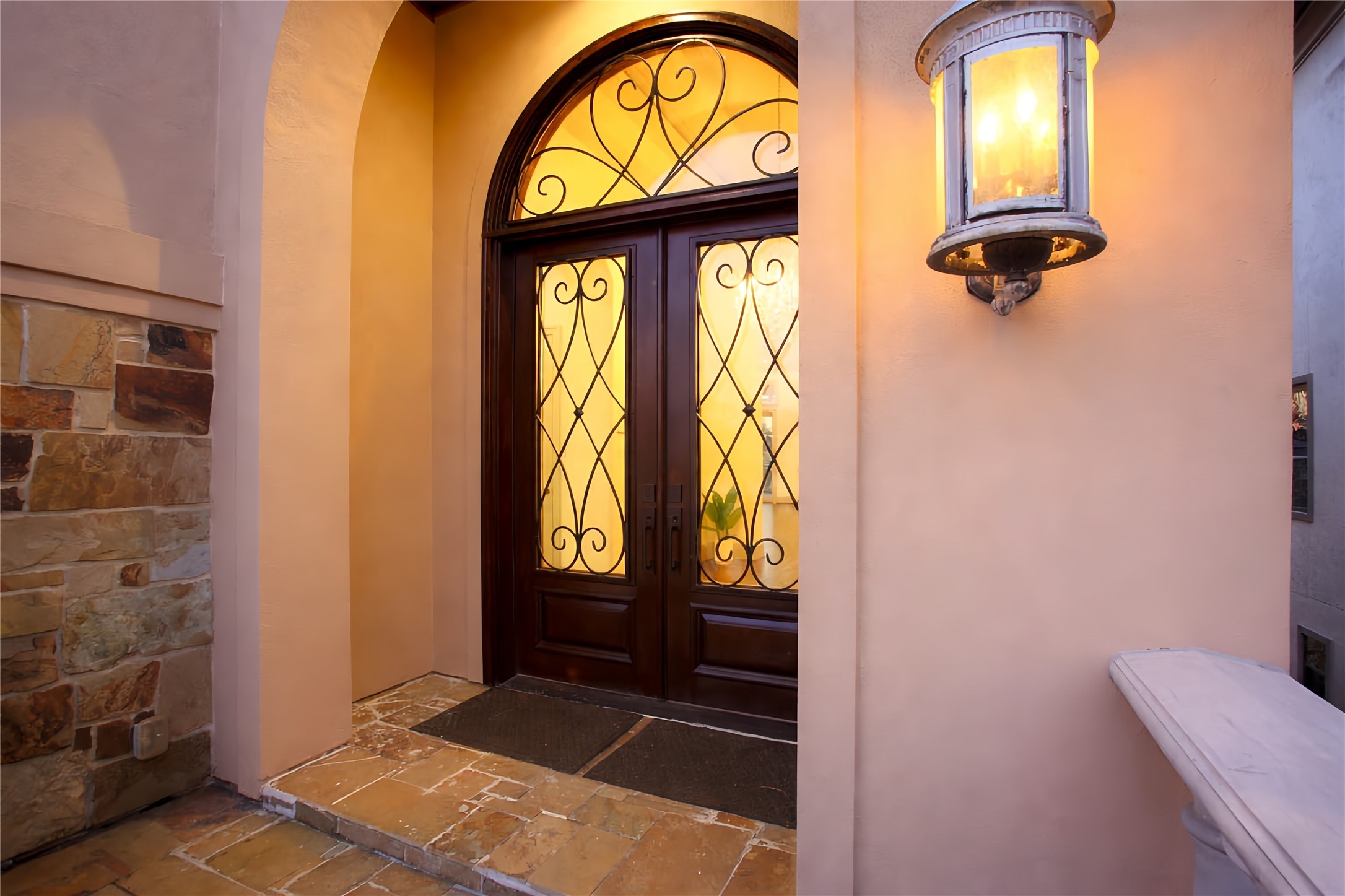 12349 Tramonto Conroe, TX 77304 - Photo 7 of 50 The entryway come with a grand wooden double door featuring intricate wrought iron designs. Warm lighting from a classic wall-mounted lantern enhances the inviting atmosphere, while the stone accents and tiled floor add a touch of luxury.