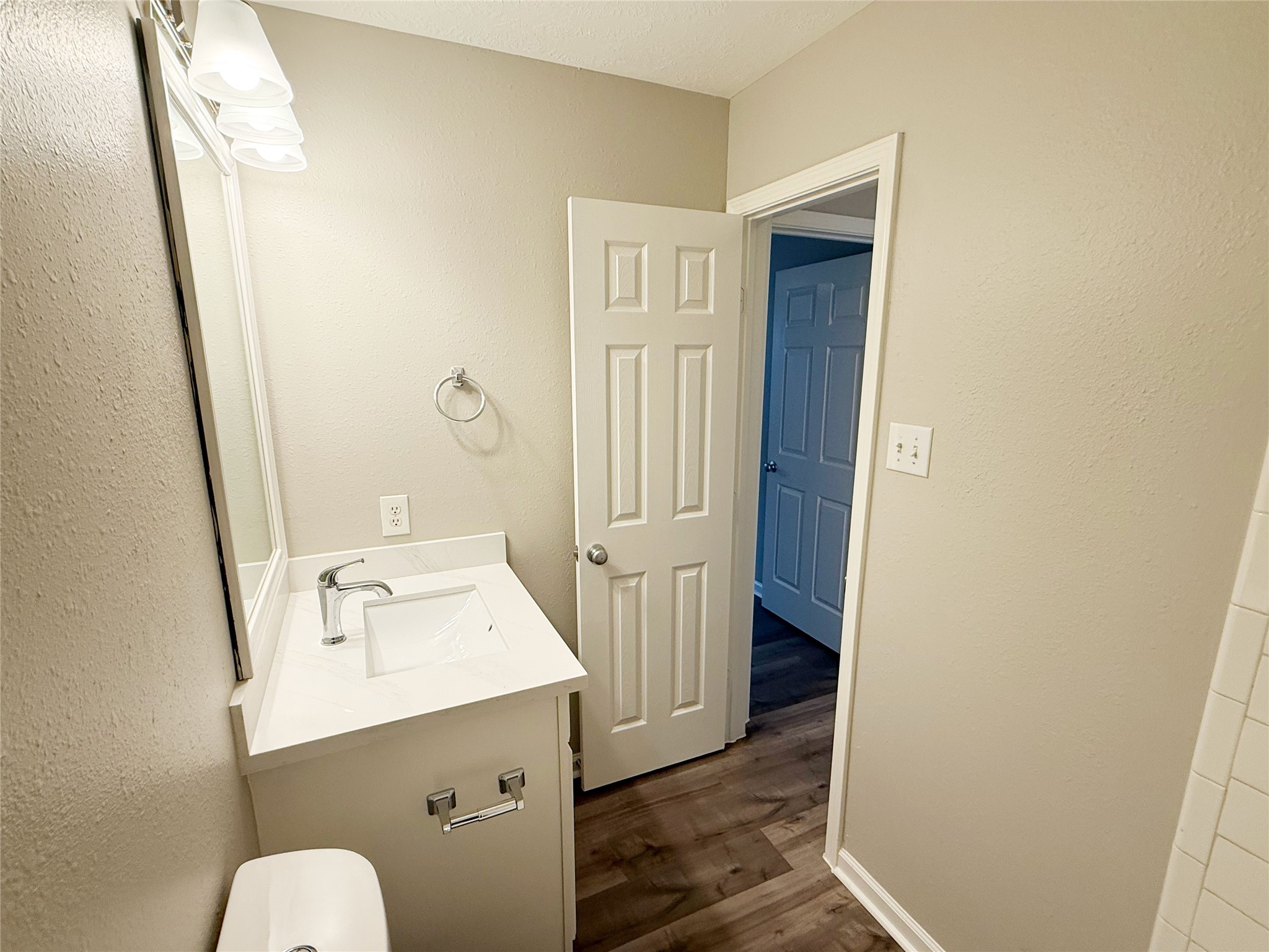 13210 Barton Meadow Lane Rosharon, TX 77583 - Photo 16 of 21 This photo shows a compact, modern bathroom with neutral-colored walls. It features a white vanity with a sleek sink, chrome faucet, and a wall-mounted mirror. The space has wood-like flooring and a door leading to a hallway.