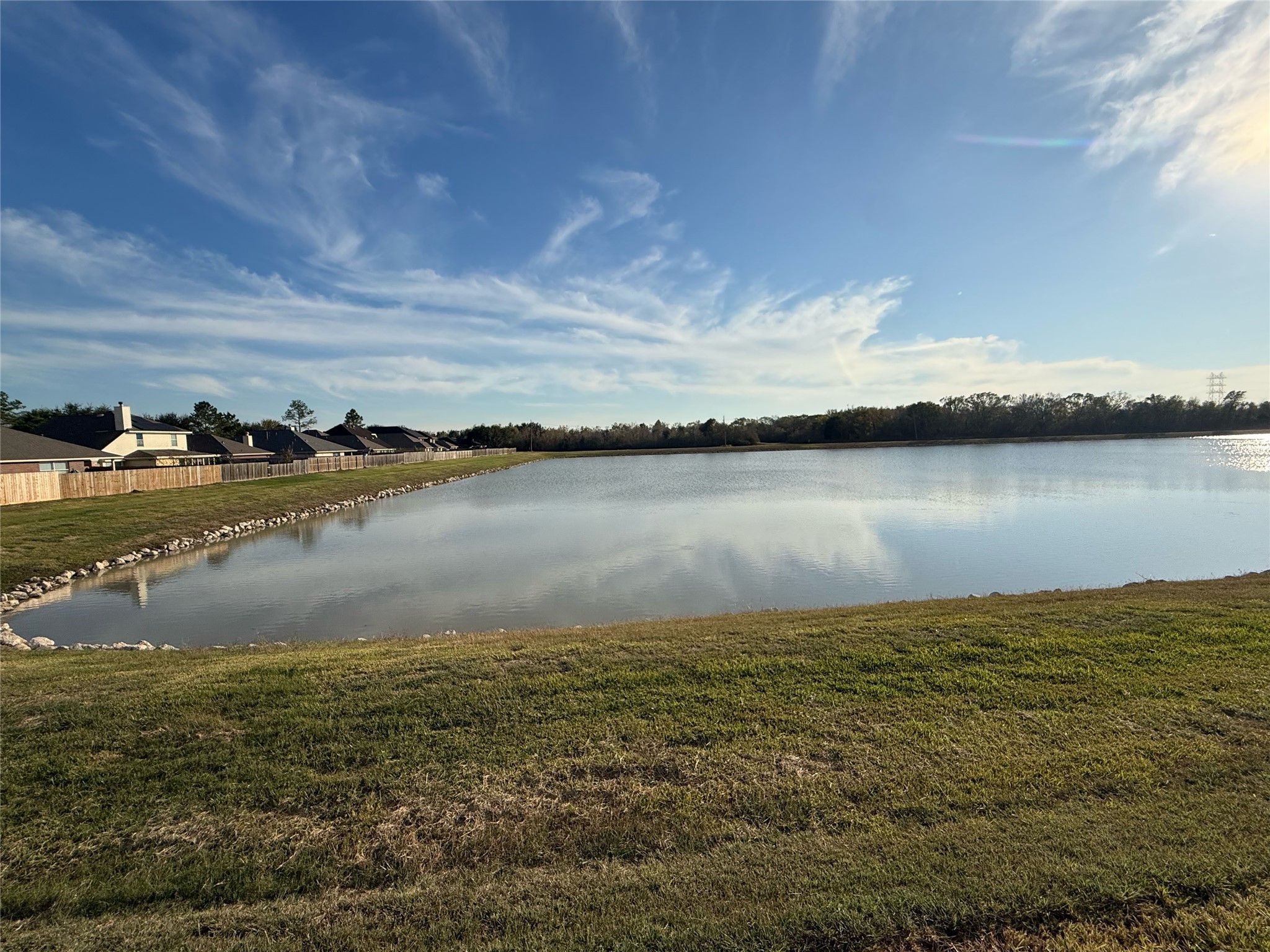 13210 Barton Meadow Lane Rosharon, TX 77583 - Photo 21 of 21 This photo showcases a serene lakeside view behind the home with calm waters, bordered by neatly maintained lawns and a row of houses in the distance, offering a tranquil and picturesque setting for a home.