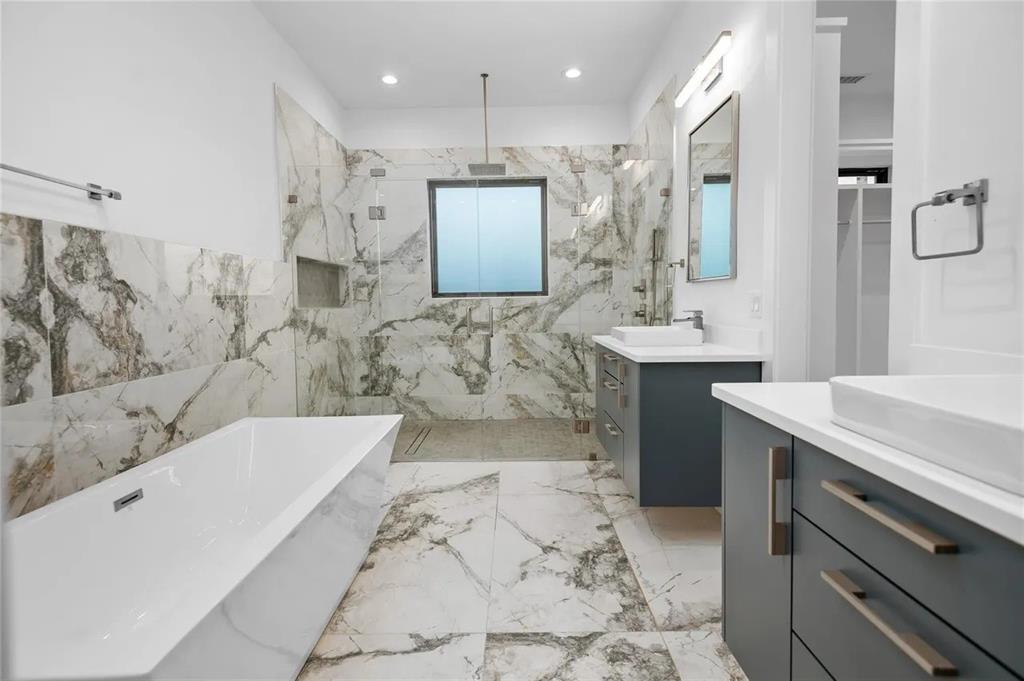 309 Kaye Street Coppell, TX 75019 - Photo 20 of 36 Spacious bathroom featuring large format marble-look tile flooring and walls, a freestanding soaking tub, a frameless glass shower enclosure, and dual vanity sinks with dark cabinetry