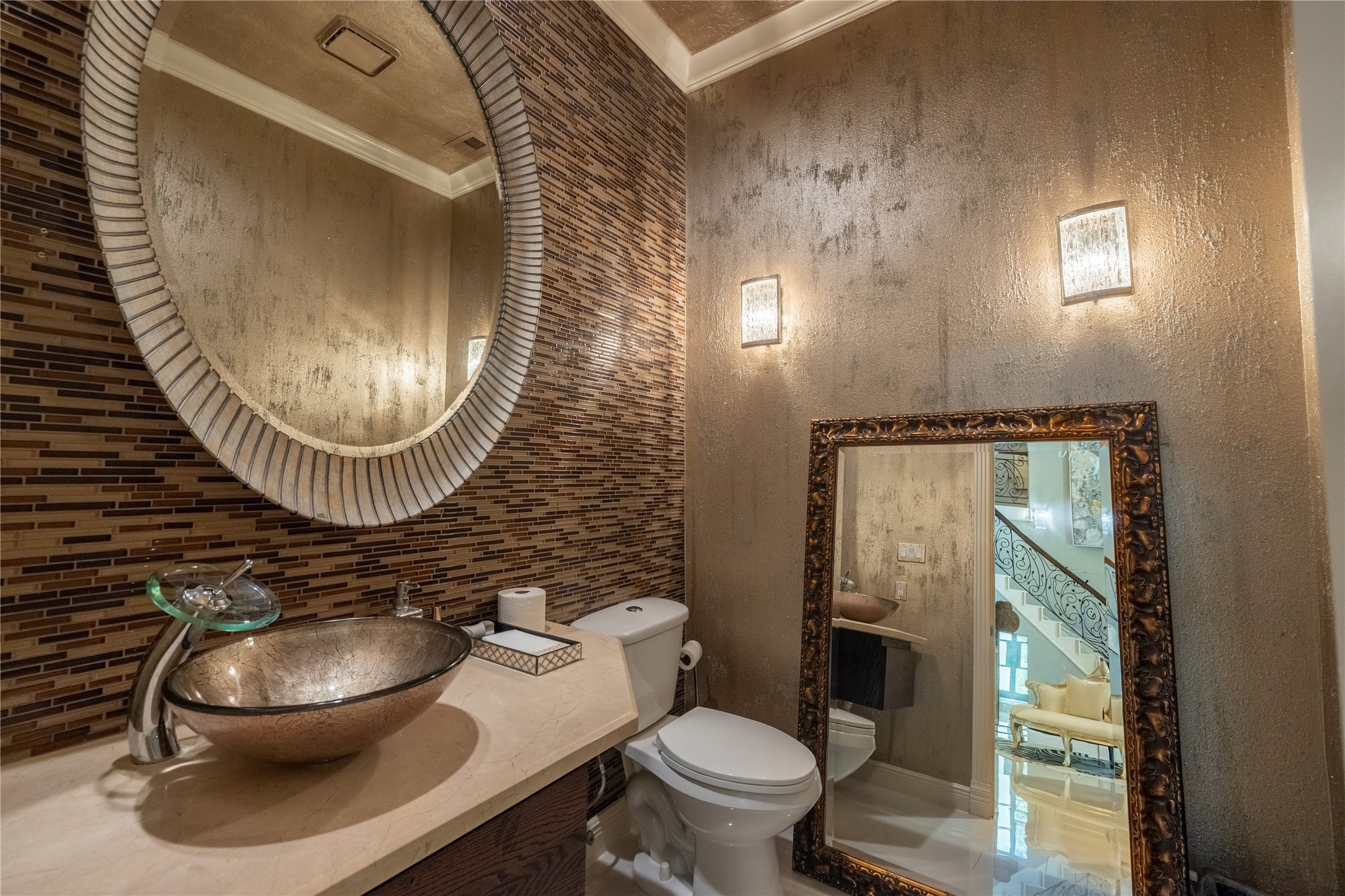 4002 Silver Ridge Boulevard Missouri City, TX 77459 - Photo 12 of 50 This is an elegantly appointed powder room featuring a textured accent wall with horizontal tile, a vessel sink atop a cream-colored vanity, and decorative mirrors. It's illuminated by stylish wall sconces and includes a modern toilet.