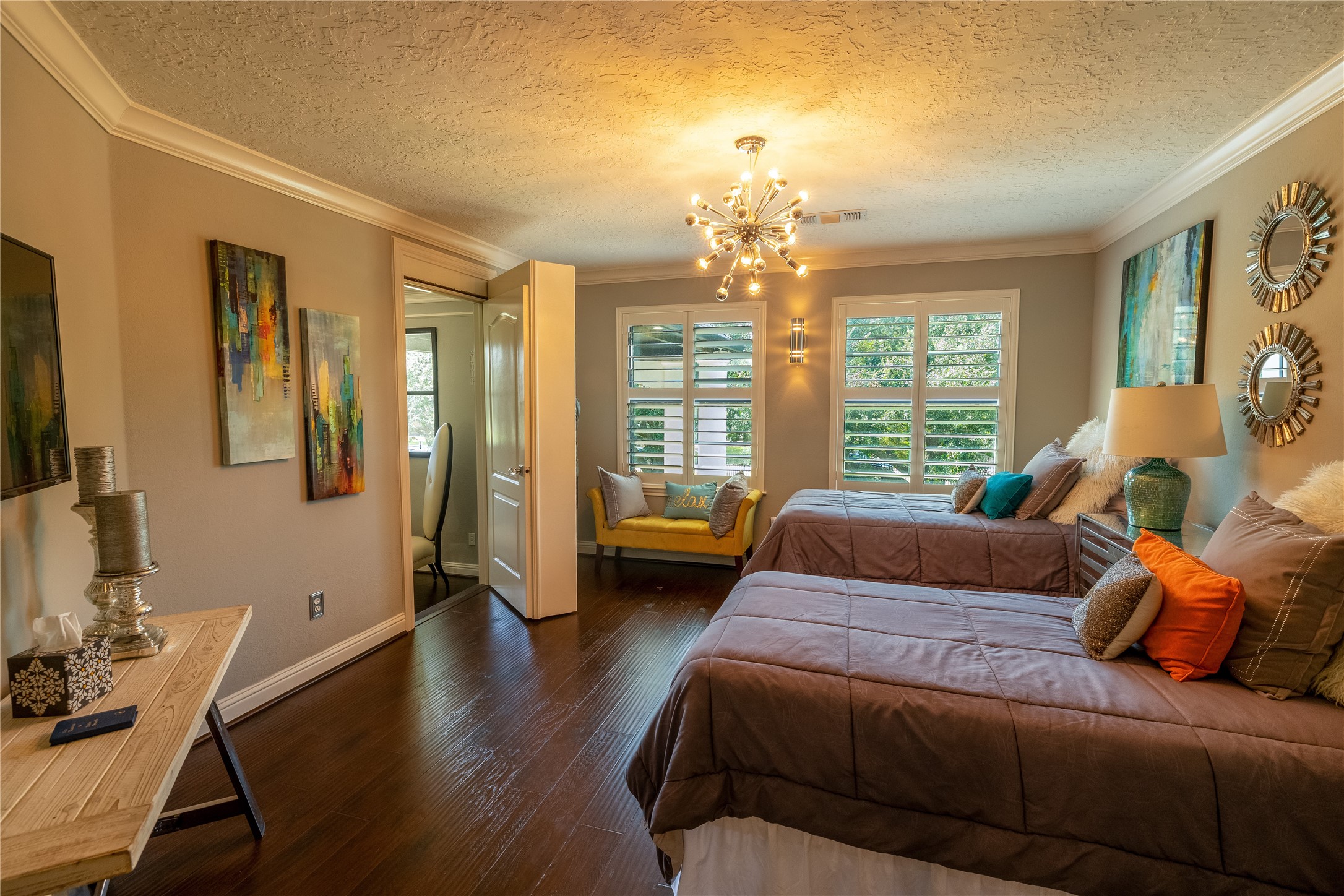 4002 Silver Ridge Boulevard Missouri City, TX 77459 - Photo 21 of 50 This photo showcases a cozy bedroom with hardwood floors, neutral walls, and textured ceiling. It features a modern chandelier, stylish decor, and a view of greenery through the window shutters. A small adjoining room with a desk suggests a private work or seating area.