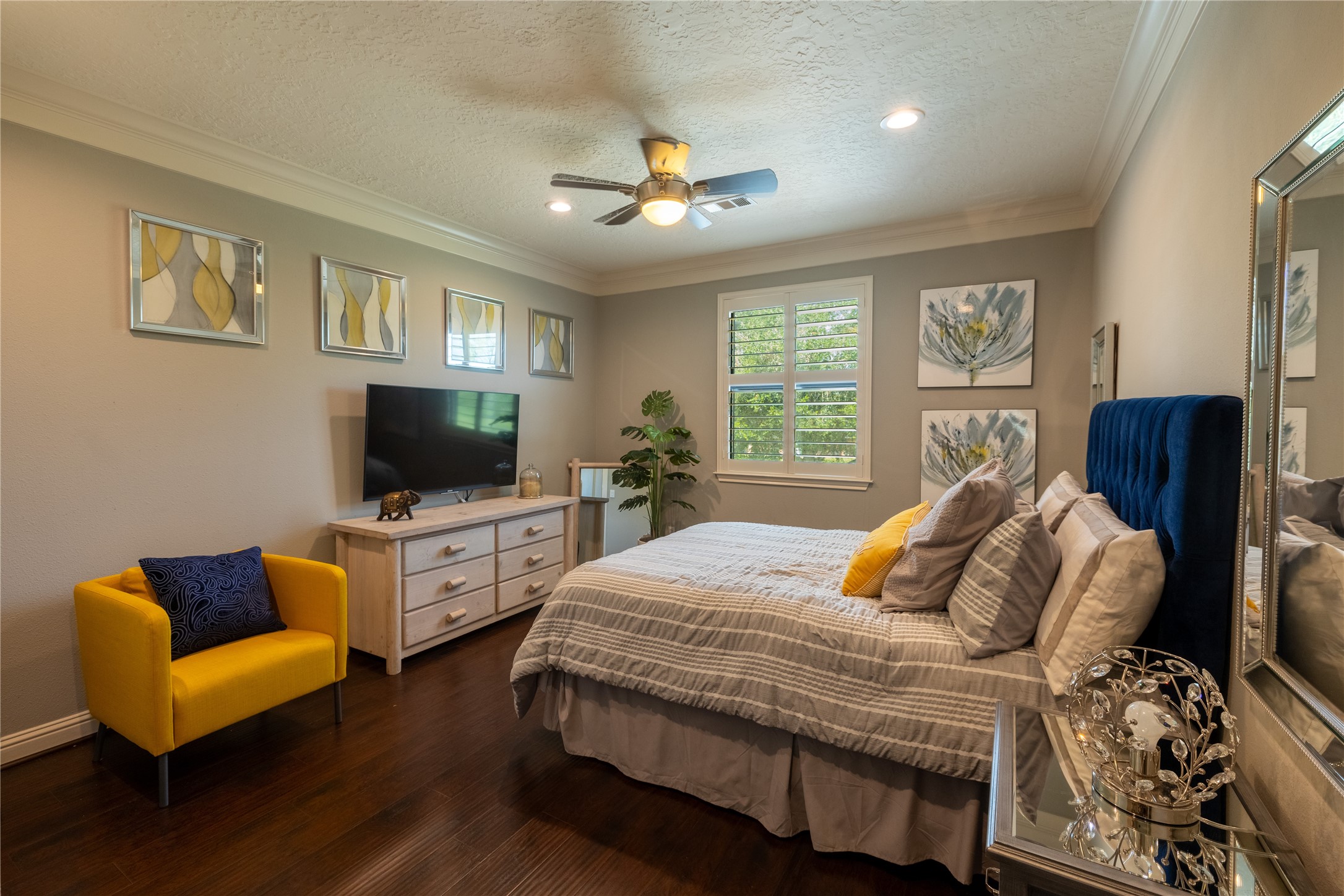 4002 Silver Ridge Boulevard Missouri City, TX 77459 - Photo 24 of 50 This is a cozy and well-appointed bedroom featuring dark hardwood floors, a ceiling fan, and natural light from windows. It has a modern aesthetic with a comfortable bed, decorative art pieces on the walls, and a vibrant yellow accent chair that adds a pop of color.