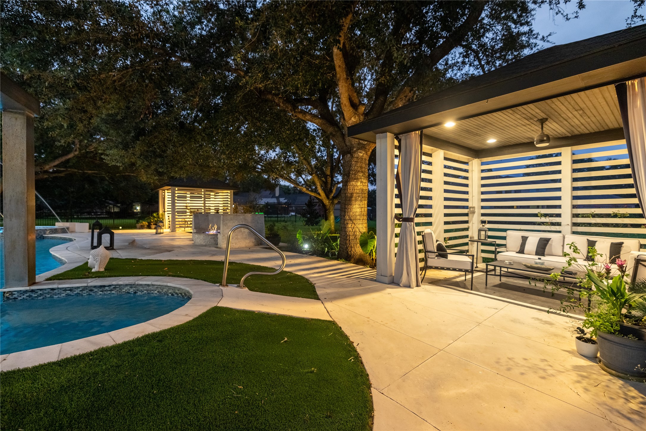 4002 Silver Ridge Boulevard Missouri City, TX 77459 - Photo 40 of 50 This is a beautifully lit, spacious outdoor area featuring a covered patio with comfortable seating, an inviting pool with a curved design, and an attractive, modern fire feature. The space is complemented by well-maintained landscaping and mature trees, offering a perfect blend of relaxation and entertainment options.